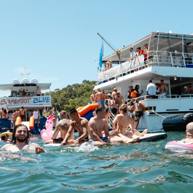 People enjoying a sunny day on the water near two docked boats, "Barefoot Blue" and another boat. Many are swimming, floating on inflatable pool toys, and socializing in groups. An enthusiastic atmosphere prevails with trees visible in the background.