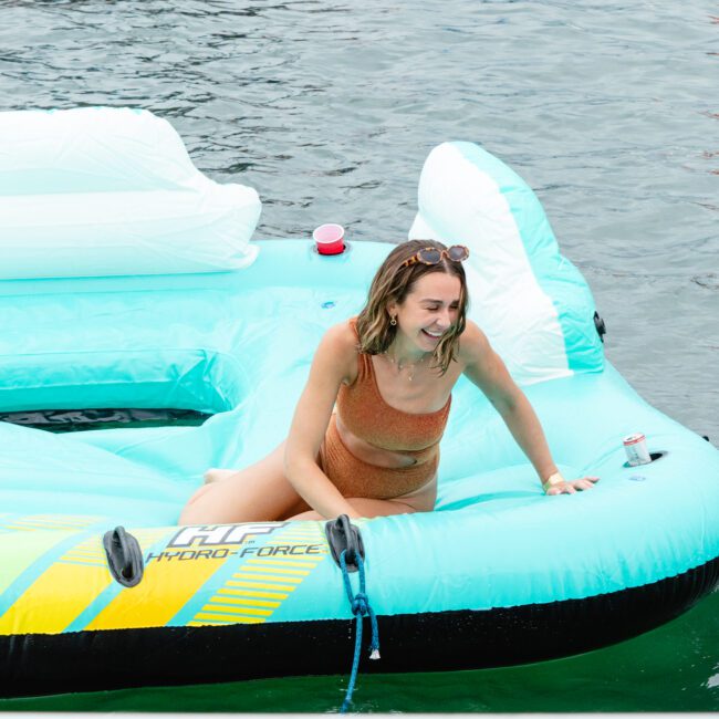 A woman in an orange bikini is smiling and climbing onto an inflatable pool float in the water. The turquoise float with white seatbacks and a cup holder holding a drink complements her sunny vibe. She has sunglasses on her head as the water ripples gently around her.