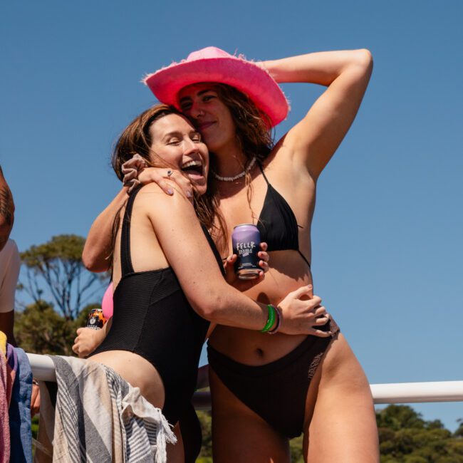 Two people in black swimsuits pose together on a boat; one person wears a pink cowboy hat and holds a can, smiling and embracing the other person. It's a sunny day with trees in the background, perfect for a private yacht charter on Sydney Harbour.