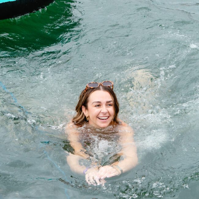 A woman with shoulder-length hair and sunglasses perched on her head is swimming in a large body of water. She looks happy and relaxed, with her arms riding atop an inflatable pink flamingo nearby. The water appears calm and inviting.