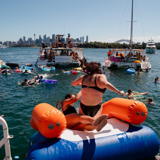 People enjoy various activities on a sunny day in a harbor, with some on boats and others swimming near colorful floats. A person jumps off an inflatable platform into the water. A luxury yacht hire in Sydney offers an exceptional way to join this vibrant scene, with the skyline visible in the distance.