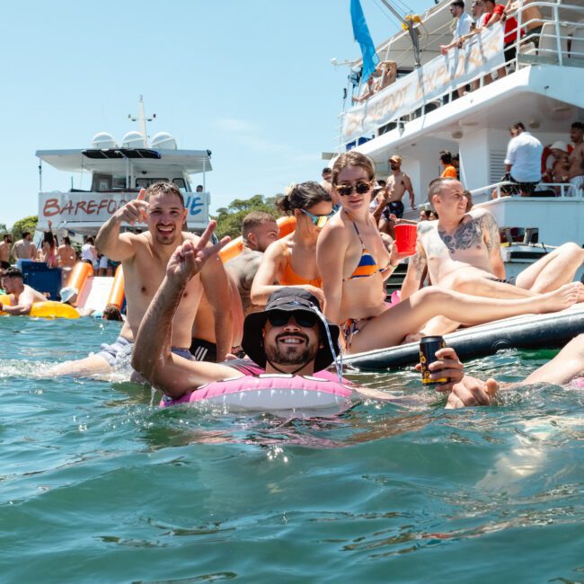 A group of people relaxing and having fun in the water near a boat. Some are floating on inflatable tubes, while others sit on the boat's deck. The scene is bright and sunny, with everyone enjoying the day. A banner on the boat reads "Barefoot Dive," adding to the festive atmosphere.
