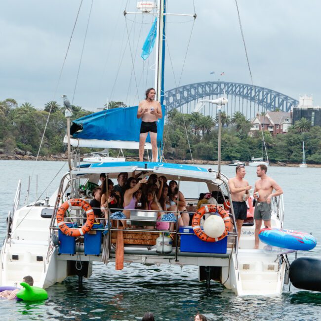 A group of friends enjoying a lively boat party on a catamaran in the harbor. Several are swimming in the water. The boat, equipped with safety rings and a blue canopy, has someone standing on the upper deck. A large bridge and lush trees create a stunning backdrop.