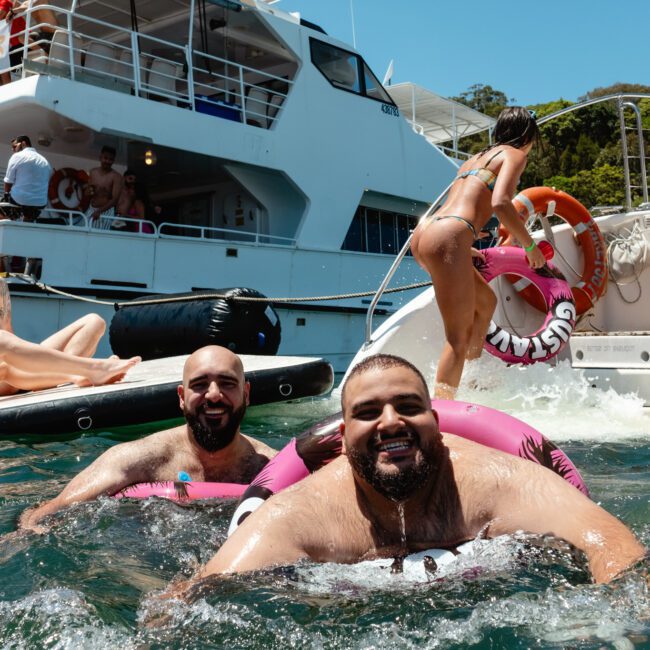 A lively scene of people enjoying a boat party. Two men smile in the water, floating on pink inflatable rings. Several people are on the boats, with some sunbathing while others socialize under a clear blue sky, against a scenic green landscape. The atmosphere feels like Instagram come to life.