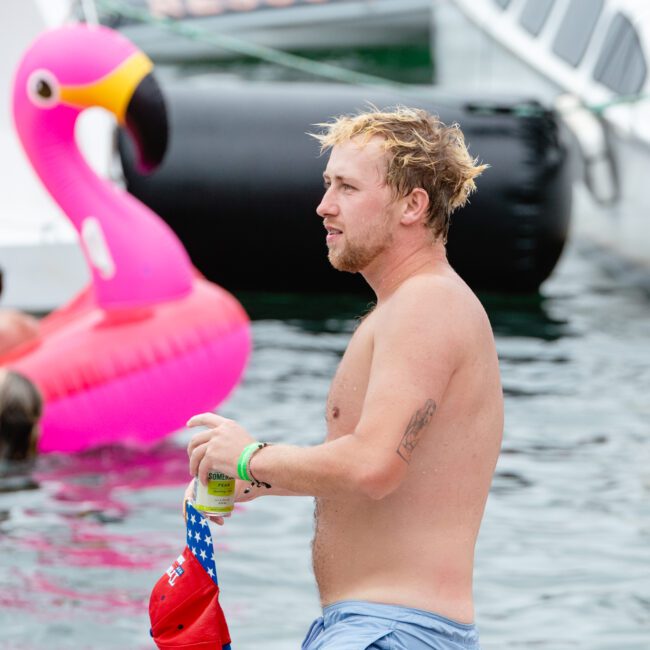 A man with wet hair stands waist-deep in water holding a beverage and a red hat with stars. He is wearing light blue swim shorts. Behind him are inflatable floats, including a large pink flamingo, paddleboards, and boats. Other people are in the background enjoying the water.