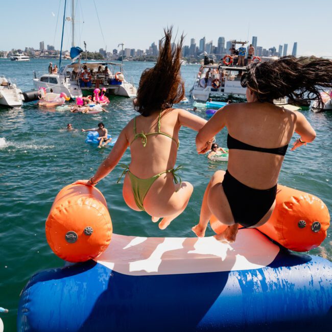 Two people in swimsuits jump off a boat onto a water inflatable in a harbor filled with other boats and swimmers. A city skyline is visible in the background, showcasing the excitement of Sydney boat party hire.