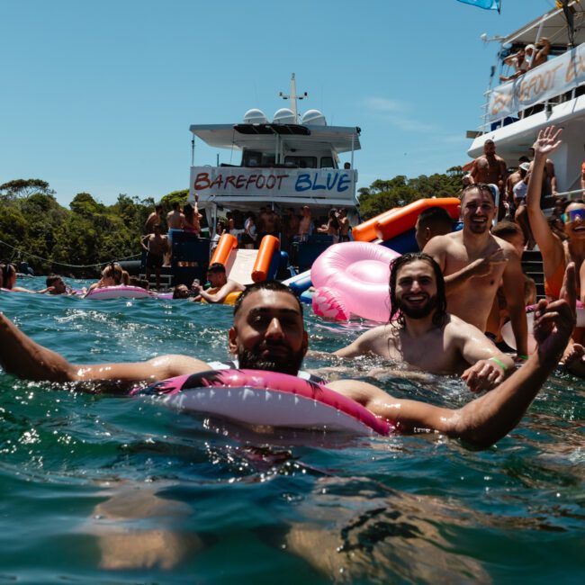 People enjoying a lively beach party on the water near boats. Some are swimming, while others relax on pink inflatables. A man in the foreground is smiling and gesturing a peace sign. In the background, there's a banner reading "Barefoot Blue" on a boat.