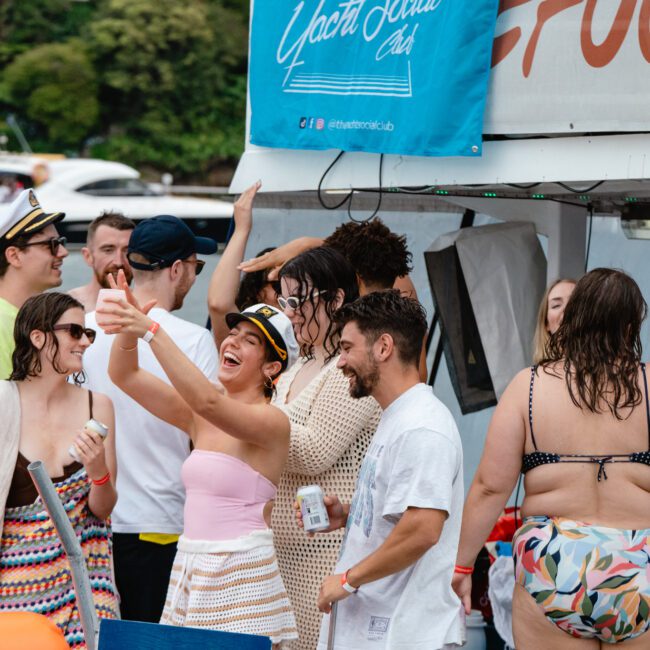 A lively group gathers on a boat for a social event. Some take selfies while others chat and hold drinks. Dressed in casual summer clothing and swimwear, they celebrate under a banner reading "Yacht Social Club." Several boats are visible in the background, adding to the festive atmosphere.