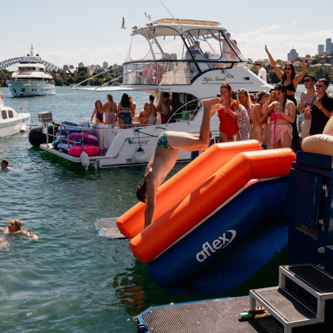 A person is diving off an inflatable water slide into a body of water surrounded by luxurious yachts. Several people are on the boats, some standing and others sitting, with a stunning cityscape visible in the background, showcasing the allure of private yacht charters in Sydney Harbour.