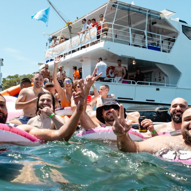 A group of cheerful people wearing swimsuits are enjoying themselves in the water near a yacht named "Barefoot Blue." The yacht is crowded with other partygoers. Some people in the water are holding up their hands in peace signs, smiling, and splashing around near the shoreline.