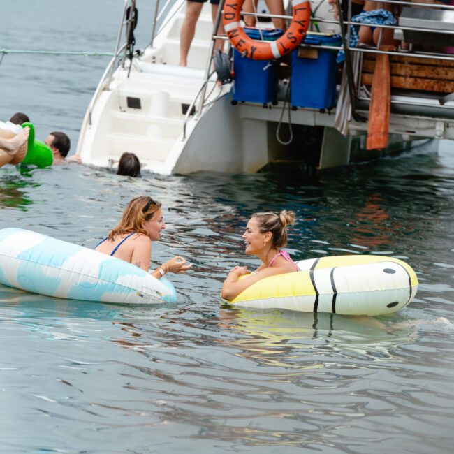 Two women relax on inflatable tubes in the water near a boat. One woman is on a blue and white tube, and the other is on a yellow and white tube. Several people enjoy their time on the boat, while others swim around. A set of steps descends from the boat to the water.