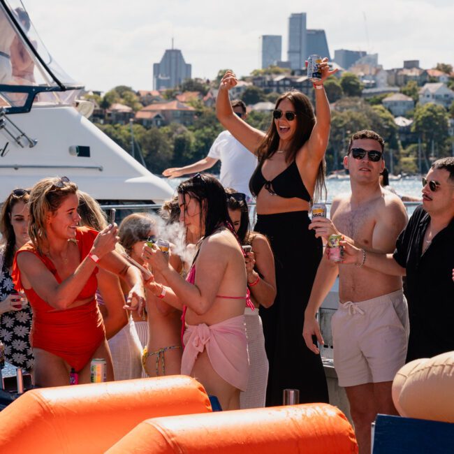 A group of people enjoying a corporate boat event near the shore. One woman in a black outfit is standing and raising her arms, while others around her are engaged in conversation and activities. Hire a luxury yacht in Sydney for your next celebration.