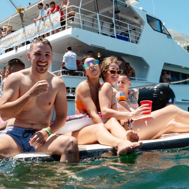 A group of people are sitting on the edge of a floating platform in the water, smiling and enjoying themselves with inflatable pool floats and toys nearby. Behind them is a large boat with more people on board, some looking out over the railing. The scene radiates joy and relaxation.