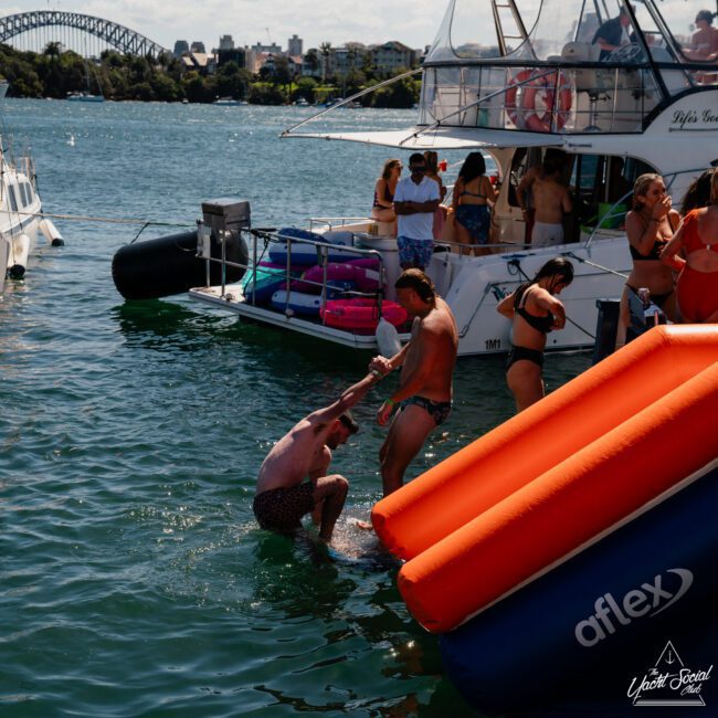 People enjoying a sunny day on a docked boat, with some swimming and using an inflatable slide to enter the water. The Sydney Harbour Bridge is visible in the background, making it perfect for a catamaran party Sydney or seamless Sydney boat party hire.