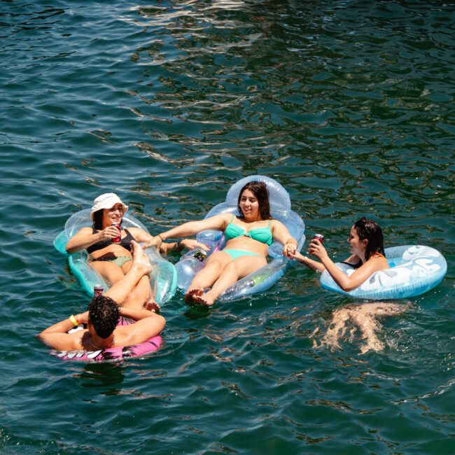 Four individuals relax on inflatable rafts in a body of water. Three of them are wearing bikinis and sunglasses, while the fourth person is in a swimsuit. The deep blue-green water enhances the scene with its tranquility, and a boat with a logo floats peacefully in the background.