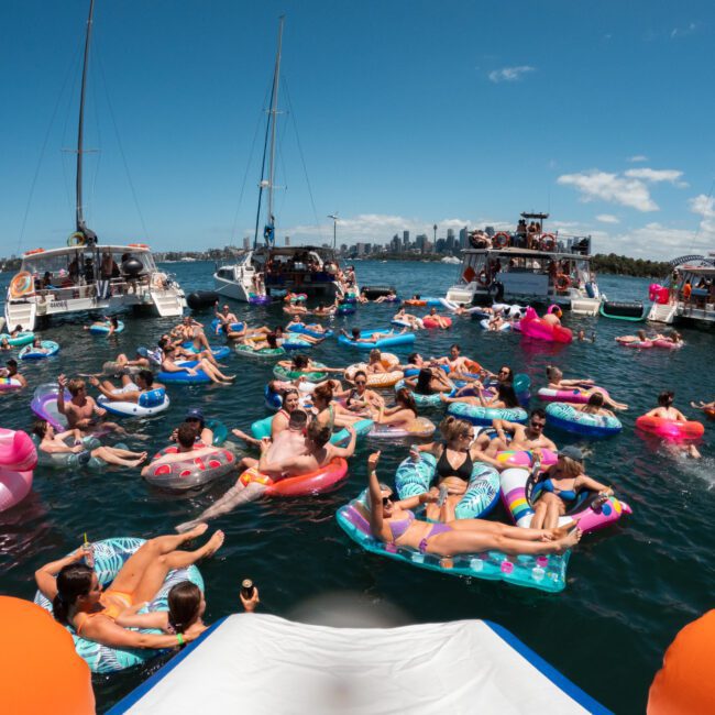 A fun gathering on the water featuring numerous people relaxing on colorful floaties in a calm body of water. Surrounding them are several boats with music playing, and in the distance, a city skyline can be seen under a clear sky. The atmosphere is joyful and festive.