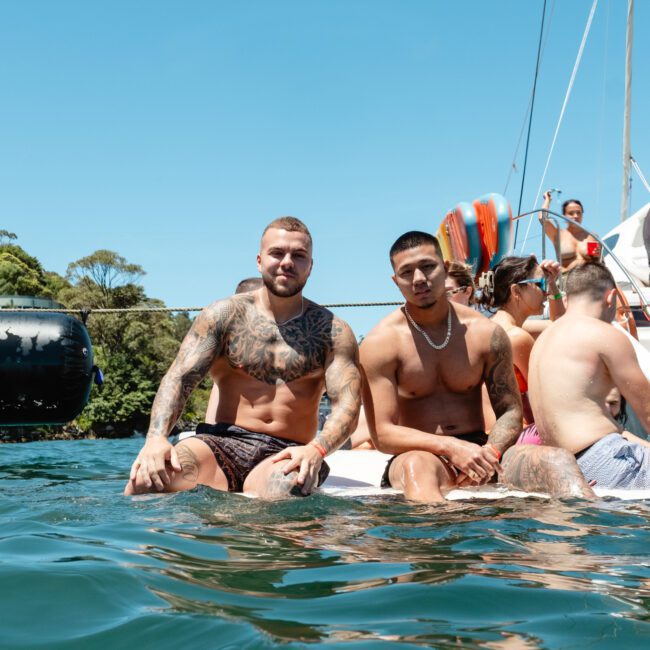 A group of people are sitting on a raft in the water near a boat. The scene appears to be a sunny day with clear skies, likely during a social gathering or vacation. Two shirtless individuals in the forefront are displaying vibrant tattoos. Trees and hills can be seen in the background.