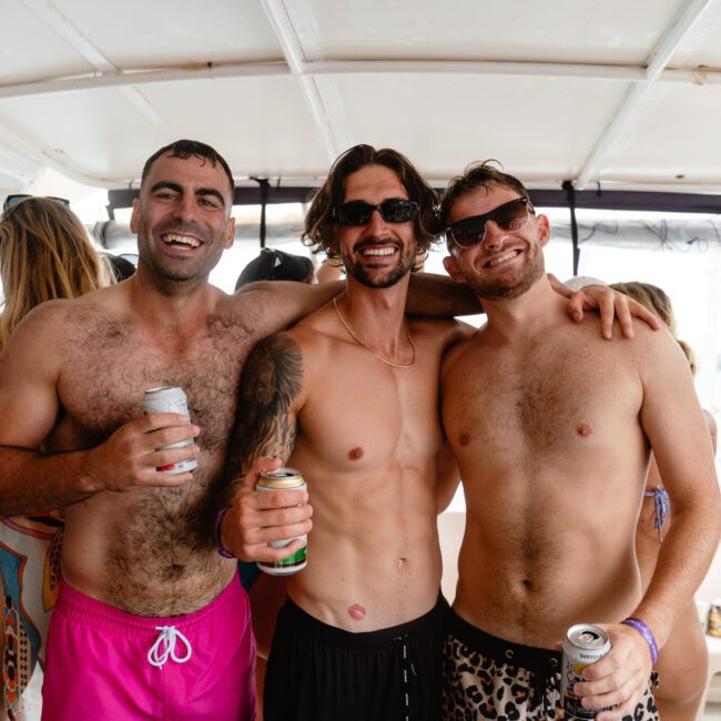 Three men stand close together on a boat, smiling and holding drinks. They are shirtless, wearing swim shorts, and sunglasses, embodying the carefree vibe of summer. Several other people socialize in the background. The atmosphere is lively and sunny, suggesting an enjoyable boat party.