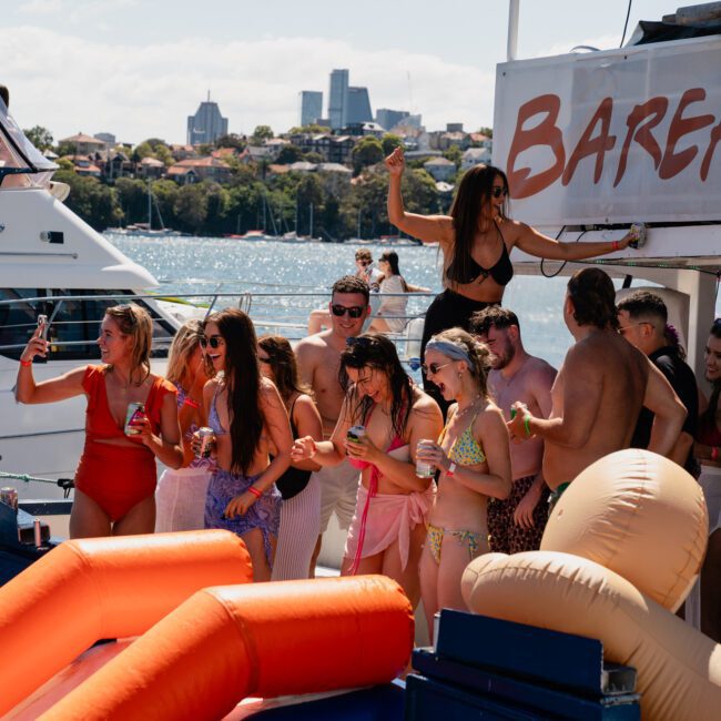 A group of people in swimsuits are partying on a luxury yacht named "Barefoot." Some are dancing, and others are holding drinks with another boat and a city skyline in the background. Ideal for private yacht charters or Sydney boat party hire, it promises an unforgettable experience on Sydney Harbour.