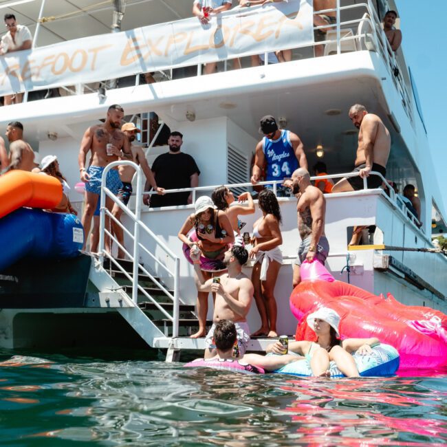 People relax and enjoy themselves on a party boat and in the water. Some are on inflatables, including a pink flamingo, while others stand or sit on the boat's deck. A waterslide extends from the "Safe-Boat Explorer" into the clear blue water, adding to the fun.