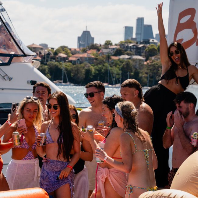Group of people in swimwear on a boat under sunny skies, enjoying drinks and celebrating. Some are posing and smiling for the camera with buildings visible in the background, highlighting the joy of a private yacht charter Sydney Harbour experience.