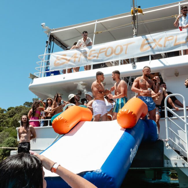A lively group of people on a boat named Barefoot Explorer enjoying a sunny day. Some are standing on a paddleboard attached to the boat's side, preparing to slide into the water, while others watch or relax on the boat's deck. Trees are visible in the background.