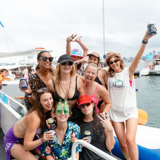 A group of nine women are on a boat, enjoying drinks and posing for the camera. They are dressed in casual summer attire with some wearing sunglasses and hats. The background shows other boats against a cloudy sky. Everyone is smiling and seems to be having a wonderful time, creating unforgettable memories.