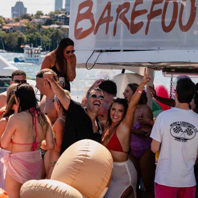 A group of people in swimwear celebrate on a boat named "Barefoot," with some raising their arms cheerfully. Sunlit waters and the city skyline are visible in the background, creating the perfect setting for a Sydney boat party hire or corporate boat events Sydney.
