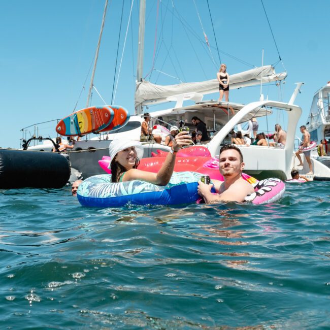 A lively scene of people enjoying a sunny day on a lake, with two individuals floating nearby on colorful inflatables. They are surrounded by several yachts and other people swimming or lounging on the boats. The festive atmosphere is enhanced by clear blue skies, calm water, and glittering reflections.