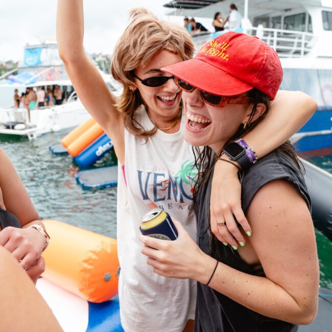 Two women smiling and laughing while standing on a dock by the water. One woman, wearing sunglasses and a tank top, has her arm around the other woman in a red cap, sunglasses, and holding a refreshing canned beverage. Boats are visible in the scenic background.