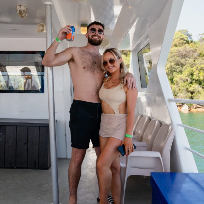 A man and woman pose on a private yacht charter in Sydney Harbour. The man is shirtless, holding a drink, and the woman is wearing a light-colored swimsuit. Trees and water are visible in the background.
