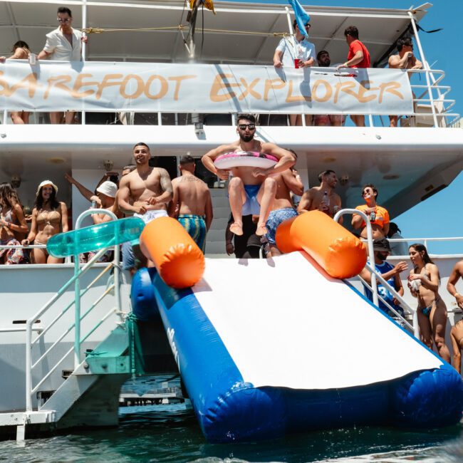 A group of people are having fun on a boat named "Barefoot Explorer." A man in an inflatable flamingo is sliding down a blue and white slide attached to the boat. Others are swimming, standing, or sitting on deck, all soaking up the sunny day on the water together.