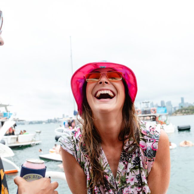 A woman wearing a pink hat and sunglasses smiles joyfully at the camera. She is on a boat with other people and yachts visible in the background, suggesting a lively and cheerful day on the water.