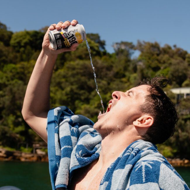 A person tilts a can to pour a drink into their mouth outdoors, with a blue towel draped over their shoulders. Trees and water are visible in the background on a sunny day, evoking the laid-back vibes of a luxury yacht hire Sydney adventure.