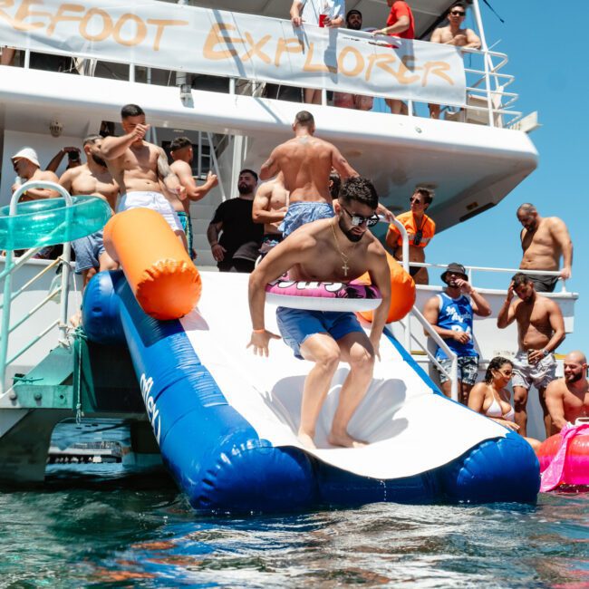 A man slides down an inflatable slide from a boat into the water, surrounded by people in swimsuits enjoying the sunny day. Some are on the boat, and others are in the water, creating a lively and fun atmosphere. The boat's name "Barefoot Traveler" is partially visible as laughter fills the air.