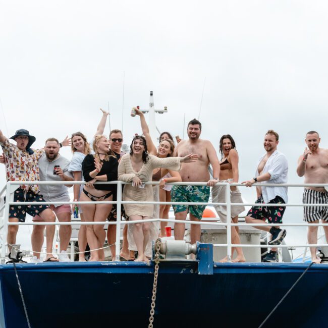 A group of people stand on the deck of a yacht, smiling and posing for the camera. They are dressed in casual summer attire, some holding drinks. Other boats are visible in the background on a cloudy day. The atmosphere appears festive and joyful as they enjoy their time together.