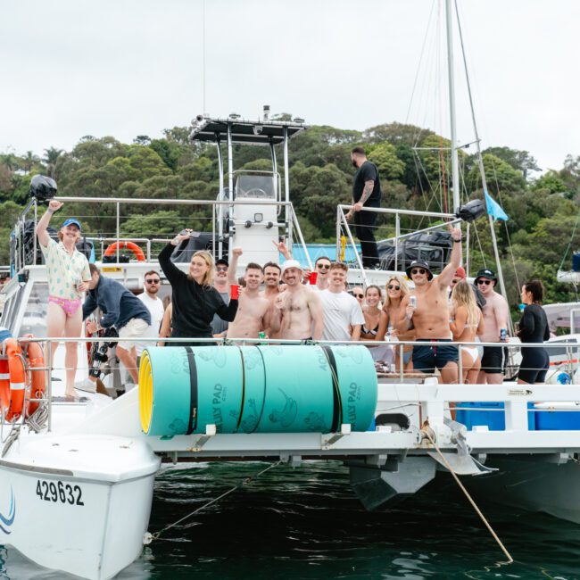 A group of people are gathered on a large boat, some in swimsuits and others in casual clothing. They are smiling and posing for the camera against a backdrop of greenery and cloudy skies. The boat has large barrels, fishing gear, and other equipment on it.