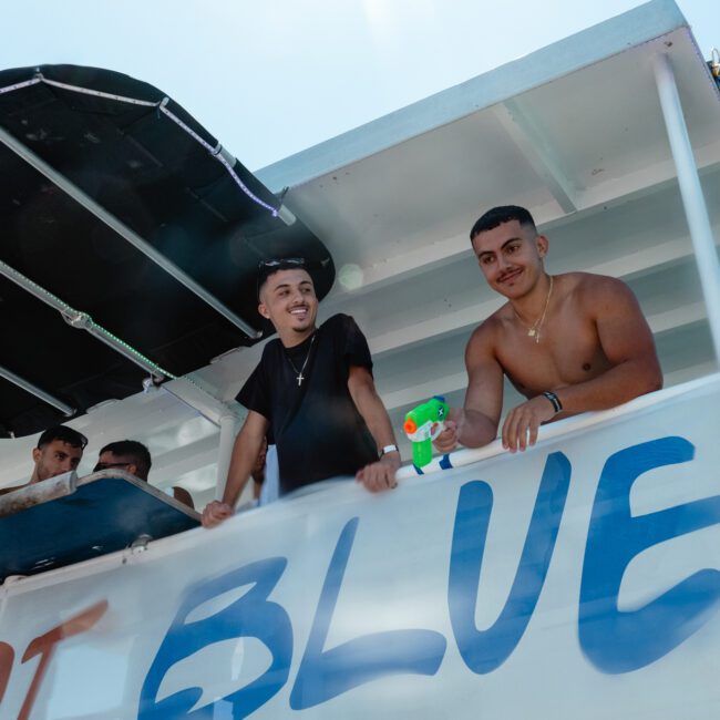 A group of young men on a boat, with one holding a colorful toy figure. The boat has a partial canopy and the word "BLUE" visibly written on its side. They appear to be enjoying themselves under the dazzling sun, surrounded by turquoise waters. The atmosphere is lively and cheerful.