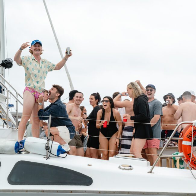 A lively group of people are gathered on a boat named "Run Runner." Some are holding drinks, while others are chatting and laughing. The atmosphere appears joyful and celebratory. The sky is overcast, and everyone is dressed in casual, beach-appropriate attire, enjoying reggae tunes in the background.