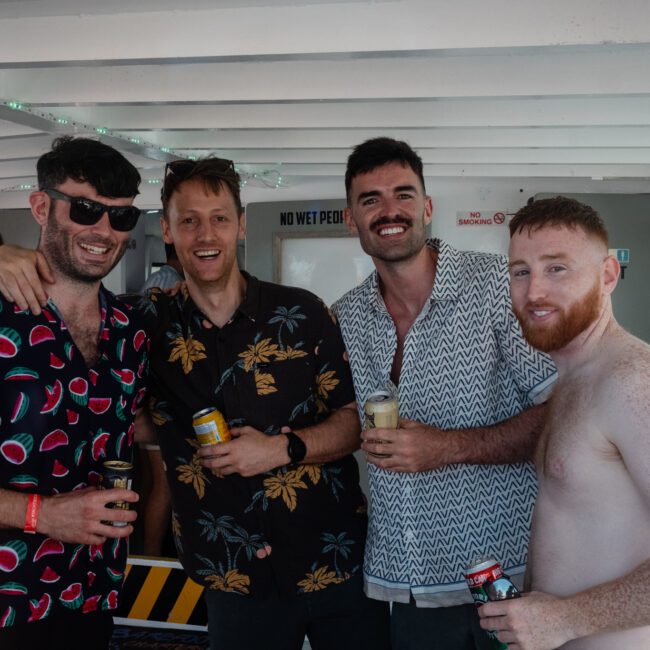 Four men are smiling and holding drinks on a boat. They are casually dressed in summer clothing, with one shirtless. Other people can be seen in the background, enjoying the vibe of a Sydney boat party hire.