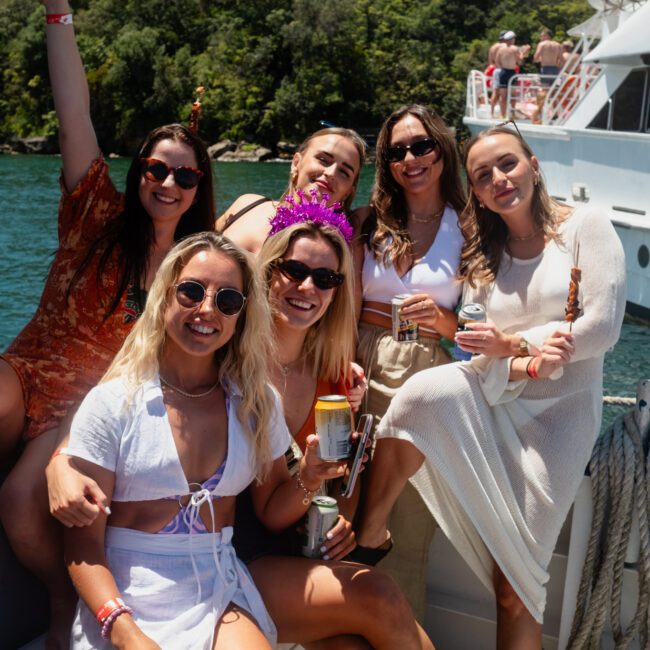 Six women are smiling and posing on a boat while enjoying a Catamaran party in Sydney Harbour. Some are holding drinks and one is wearing a purple crown. A large yacht and scenic greenery serve as the perfect backdrop.