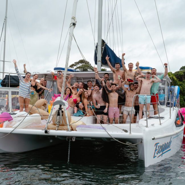 A group of people on a sailboat, all smiling, with many raising their arms in enthusiasm. The boat is docked in calm water, surrounded by other boats. In the background, lush greenery adorns the shore. The celebratory mood captures a lively and vibrant scene of joy and camaraderie.