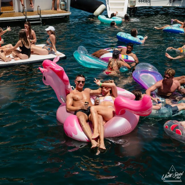 A group of people are relaxing on various inflatable floats in the water. A man and woman are lounging on a pink unicorn float, holding drinks and gesturing towards the camera. This scene captures the joy of a private yacht charter in Sydney Harbour, perfect for a luxurious day out.