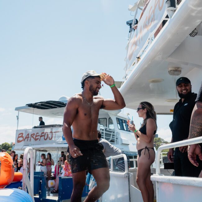 A man in a cap and shorts is walking on a boat while several people, including one taking a photo, are engaged in various activities around him. This scene captures the essence of a Catamaran party in Sydney.