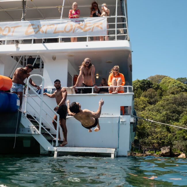 A group of people on a boat watch as a person dives backward into the water. The boat has an orange slide and "Barefoot Explorer" written on it. Trees and blue sky are in the background, perfect for your next corporate boat event in Sydney.