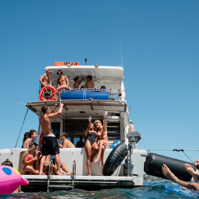 People enjoying a Catamaran party in Sydney, with some on the deck and others in the water on inflatable floats, under a clear blue sky.
