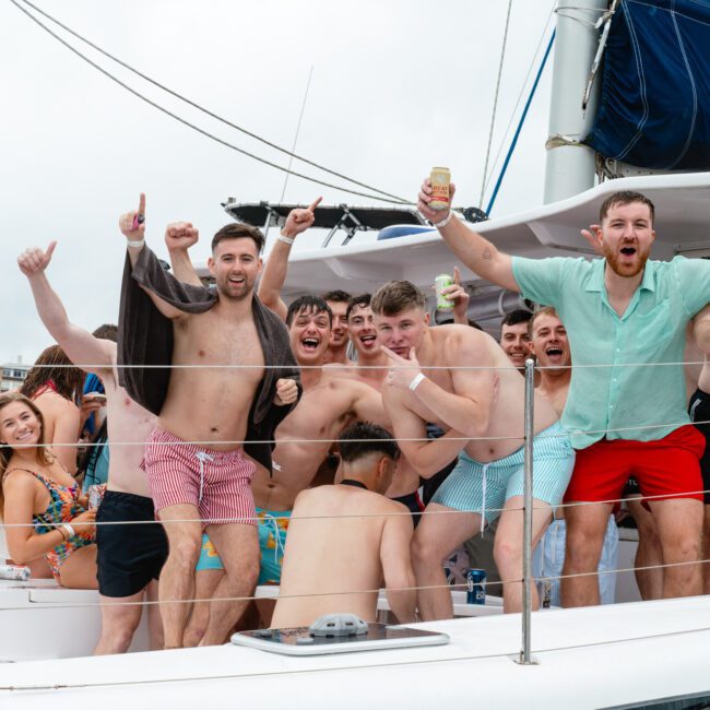 A group of cheerful people on a boat, most of them in vibrant swimwear, are celebrating and posing for the camera. They are smiling, with some holding drinks and raising their arms in excitement. The background shows part of the boat, railings, and a cloudy sky.