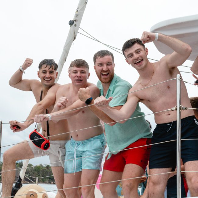 Five men are posing energetically on a boat, flexing their arms and smiling at the camera. They are dressed in casual summer attire, with some shirtless and others in swimming shorts. The background shows a cloudy sky with distant buildings and the ocean, adding to the adventurous vibe of their day.