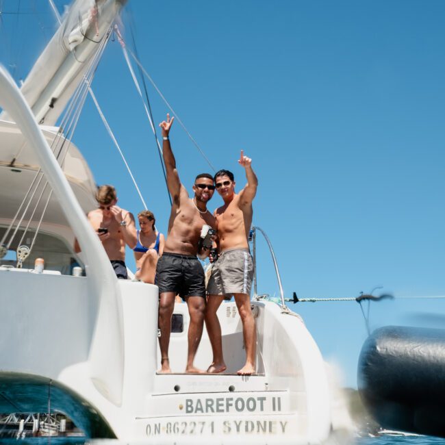 Two men in swim trunks stand on the back of a yacht named "Barefoot II" with arms raised in celebration. Other individuals are seen on the yacht, enjoying the sea in the foreground—a perfect scene for luxury yacht hire Sydney or catamaran party Sydney.