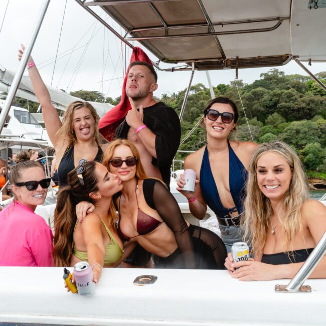 A group of people enjoying a boat party. Some are holding drinks, a few are wearing swimsuits, and they are all smiling and posing for the camera. The background features another boat and lush green trees. One person playfully licks another's face while laughter fills the air with vibrant energy.